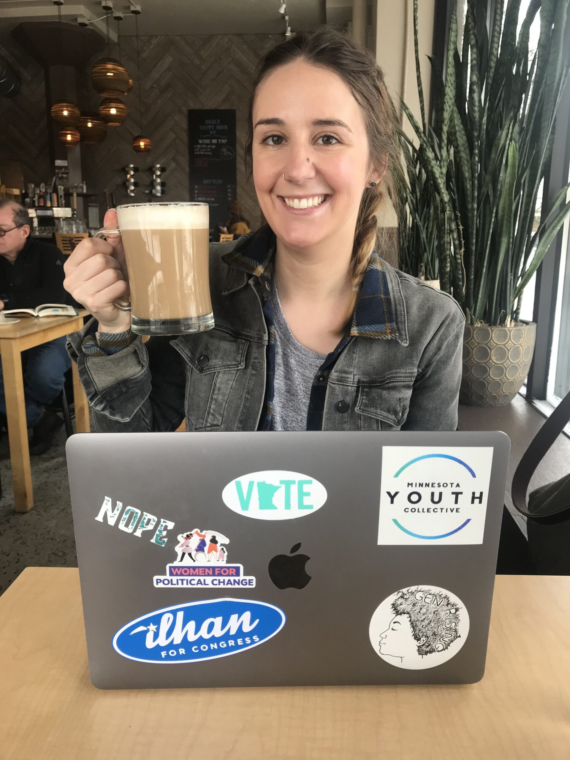 A young woman sits in a cafe, hold a coffee mug and smiles at the camera, a laptop open in front of her